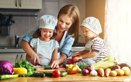 Mama i dwójka dzieci w kucharskich strojach przygotowują danie. Mama uczy córkę kroić ogórka. Na całym stole znajdują się poukładane warzywa i owo