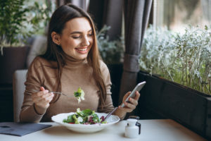 Kobieta siedząca przy stole, jedząca sałatkę, w ręku trzyma smartfona.